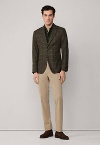 Brauner kariertes Sakko mit Revers, kombiniert mit einem dunkelgrünen Hemd und hellbeigen Hosen. Braune Stiefeletten runden das Outfit ab.