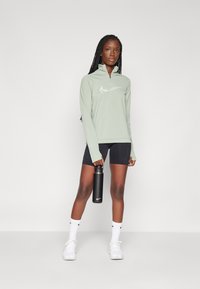 Sudadera de manga larga de color verde claro con cremallera hasta el pecho y un logo reflectante, combinada con pantalones cortos negros y zapatillas blancas. Sosteniendo una botella de agua negra.