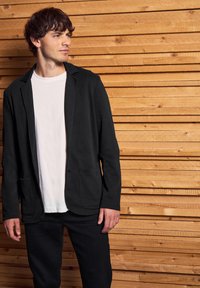 Svart vävd blazer med två framfickor, bärs över en vit t-shirt, tillsammans med svarta byxor. Mot en bakgrund av träplankor.