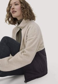 Fleecejacke mit einem hellbeigen oberen Teil und einem dunkelvioletten unteren Abschnitt, mit hohem Kragen und Reißverschluss. Strukturiertes Gewebe.