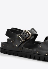 Sandali neri in pelle con dettagli in gomma dorata, cinturini regolabili con fibbie e suola testurizzata per una migliore aderenza. Design open-toe con profilo piatto.