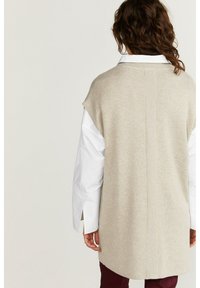 Beige stickad ärmlös tröja med rak passform och detalj i ryggen, lagd över en vit skjorta med breda manschetter och vinröda byxor.