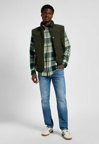 Gevoerde groene vest met geribbelde accenten, gelaagd over een zwart-groene geruite shirt, gecombineerd met lichtblauwe jeans en witte sneakers.