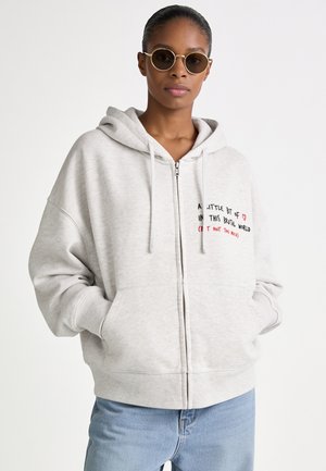 Vrouw met ronde zonnebril, lichtgrijze zip-up hoodie met tekst en lichtblauwe spijkerbroek, staand met handen in de zakken tegen een witte achtergrond.