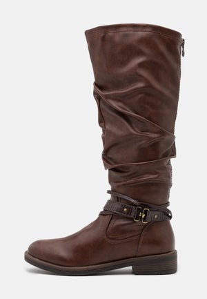 Ruskea nahkainen nilkkaa ylettyvä saapas, jossa löysä muotoilu, koristeelliset nilkkahihnat, matala kasaantunut korko ja sivusaumallinen vetoketjukiinnitys.