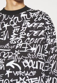 Černá mikina s bílým graffitovým textem a grafickými prvky. Má kulatý výstřih a ribbované manžety. Měkká, strukturovaná tkanina.