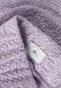 Tissu tricot violet lavande avec des paillettes argentées, présentant un bouton transparent cousu sur une boucle en tissu blanc.