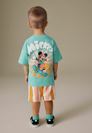 Kind mit dem Rücken zur Kamera, trägt ein türkisfarbenes Shirt mit Mickey-Mouse-Skateboardmotiv und orange-weiß gestreifte Shorts.