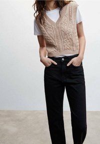 Pull sans manches en tricot à torsades beige avec col en V. Porté par-dessus un T-shirt blanc. Associé à un jean en denim noir taille haute.