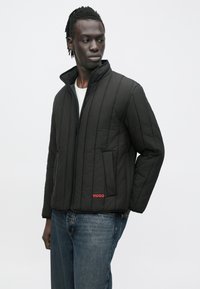 Schwarze gesteppte Jacke mit Stehkragen, Frontreißverschluss und Seitentaschen; mit rotem "HUGO"-Logo unten links.