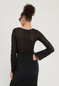 Schwarzes Spitzenoberteil mit langen Ärmeln und floralem Muster, kombiniert mit einem hoch geschnittenen schwarzen Jeansrock, der eine taillierte Silhouette und ausladende Ärmel hat.