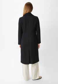 Long manteau noir avec un col, coupe droite à simple boutonnage, silhouette droite, et une fente au dos, porté sur un pantalon large de couleur claire.