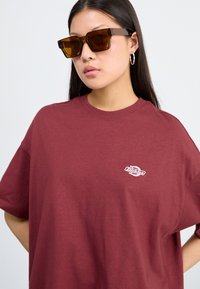 T-shirt en coton bordeaux avec une coupe décontractée et un petit logo blanc. Modèle portant des lunettes de soleil marron oversize et des boucles d'oreilles en argent.