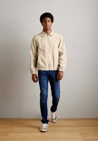 Helle beige Zip-Jacke mit elastischen Bündchen, getragen über blauen Jeans und weißen Sneakern mit grünen Akzenten, steht auf einem Holzboden.