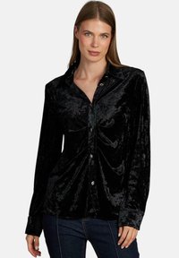 Camisa de terciopelo negro con cierre de botones en el frente, mangas largas y cuello puntiagudo. Detalles fruncidos en el busto y acentos de botones brillantes.