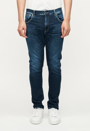 Jean en denim bleu foncé à coupe slim, avec des surpiqûres contrastantes, cinq poches et des ferrures argentées. Associé à des baskets blanches.