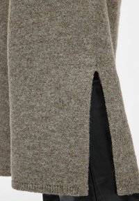 Tissu tricoté marron avec fente latérale révélant un pantalon noir en dessous, montrant la texture du vêtement et les détails de superposition.