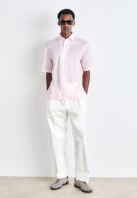 Korte mouwen, lichtroze linnen shirt met knopen aan de voorkant, gecombineerd met witte, relaxed-fit broek en grijze schoenen. Model draagt een zonnebril.