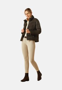Black puffer jacket with a high collar, zip closure, and side pockets. Worn over a light shirt and beige fitted leggings, paired with brown boots.