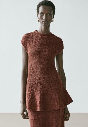 Persona alta che indossa un vestito marrone lavorato a maglia senza maniche con orlo svasato, in piedi contro uno sfondo chiaro e semplice, con orecchini a cerchio dorati.