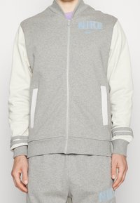 Man som bär en grå och vit Nike zip-up jacka och matchande gråa sweatpants med Nike-logotyper på bröstet och benet.