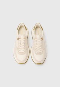 Kreemivärvi spordijalatsite paar, millel on lipsukujundus, oliivrohelised aktsendid kanna ja taldadel ning väikesed kullaga logod lipsude lähedal.