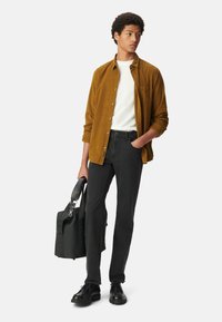 Modelo vistiendo una camisa de pana mostaza, camiseta blanca, jeans negros y zapatos negros, sosteniendo una mochila negra.