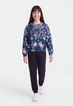 Fille souriante aux longs cheveux foncés porte un sweat-shirt bleu marine avec des motifs roses et blancs, un pantalon noir et des baskets blanches, marchant vers l'avant.