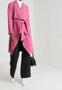 Rosa Wickeljacke mit breiten Revers und einem zum Binden Gürtel, kombiniert mit einem weißen Rollkragenpullover, schwarzen weit geschnittenen Hosen und weißen Knöchelstiefeln.