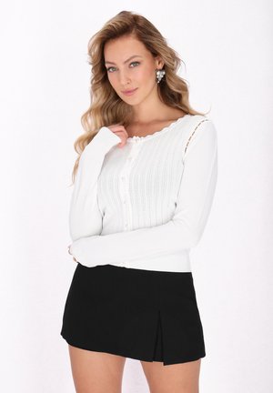 Cardigan alb din tricot cu nervuri, cu un decolteu zimțat, având nasturi pe față și o deschidere la manșetă, asortat cu o fustă mini neagră.