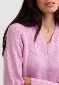 Różowy sweter z dzianiny z dekoltem w serek, prążkowaną fakturą i długimi rękawami. Cechuje się miękką tkaniną oraz subtelnym splotem na ramionach.