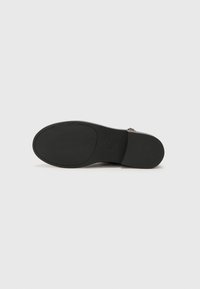 Semelle noire d’une chaussure habillée avec une zone de grip texturée et un petit logo embossé près du talon sur un fond clair uni.