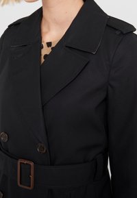 Žena v čern�ém trench coat s páskem přes béžový svetr s černými puntíky, zobrazená od hrudníku nahoru.