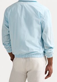 Ljusblå jacka med elastiska ärmslut och nederkant, tillverkad av ett lätt material. Bakifrån syns en enkel design utan utsmyckningar.