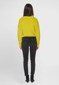 Jasně žlutý pletený svetr s volným střihem, kombinovaný s úzkými černými džíny a černými kotníkovými botami, pohled zezadu.