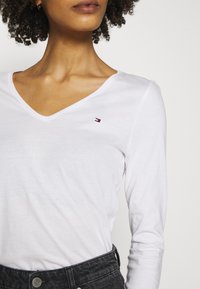 Biała, długa koszula z dekoltem w serek, z małym czerwonym, niebieskim i białym logo na lewej piersi, w połączeniu z ciemnymi dżinsami z zapięciem na guzik.