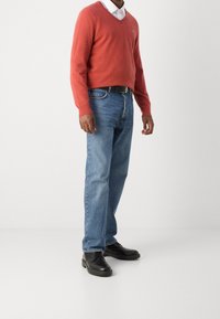 Homme portant un pull à col en v orange, une chemise blanche à col, un jean bleu avec une ceinture noire, et des chaussures noires à lacets, se tenant sur un sol blanc.