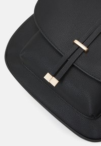 Sac à main en cuir noir texturé avec fermeture à rabat, doté de deux fines sangles et d'accents métalliques rectangulaires dorés sur un fond blanc.