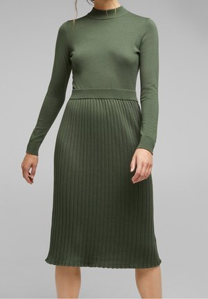 Femme portant une robe midi en maille côtelée vert olive avec manches longues et col montant, debout devant un fond gris uni.