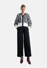 Jeune femme portant un pantalon large noir, un cardigan rayé noir et blanc boutonné, et des bottines rouges foncées, debout les mains dans les poches.