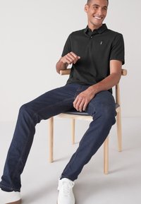 Camisa polo negra con un pequeño logo, combinada con unos jeans de color azul oscuro. El modelo está sentado en una silla de madera, vistiendo zapatillas blancas.