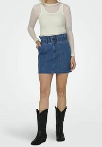 Maglietta bianca a maniche lunghe con coste abbinata a una gonna di jeans blu con cintura arricciata e tasche frontali. Indossata con stivali da cowboy neri.