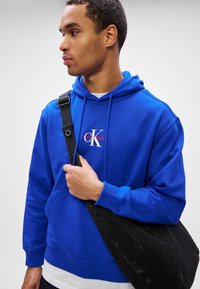 Fiatal férfi élénk kék Calvin Klein kapucnis pulóverben, egy nagy fekete válltáska pántját fogja, és balra néz.