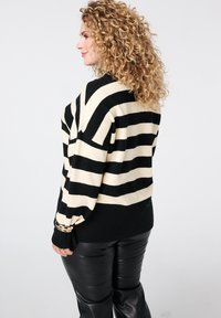 Pull rayé noir et crème à coupe décontractée, avec des poignets côtelés, une texture douce et des épaules arrondies. Associé à un pantalon noir brillant.