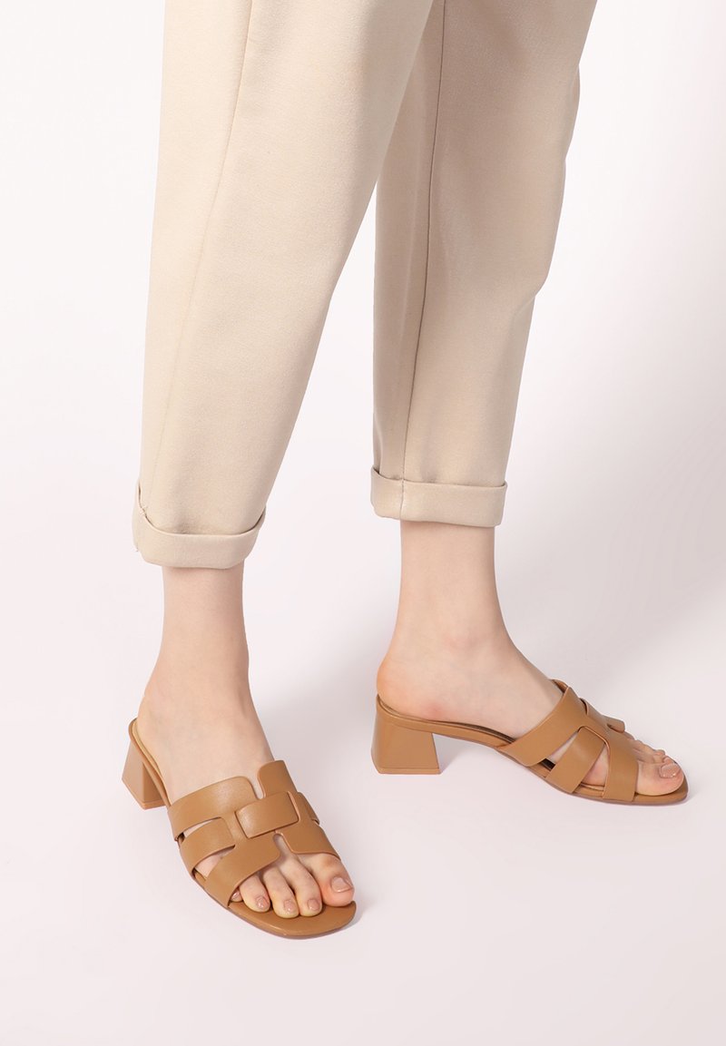 Bruine sandalen met hakken, open teen en gekruist riempje ontwerp. Gemaakt van een glad materiaal, met een blokhak en een platte zool.