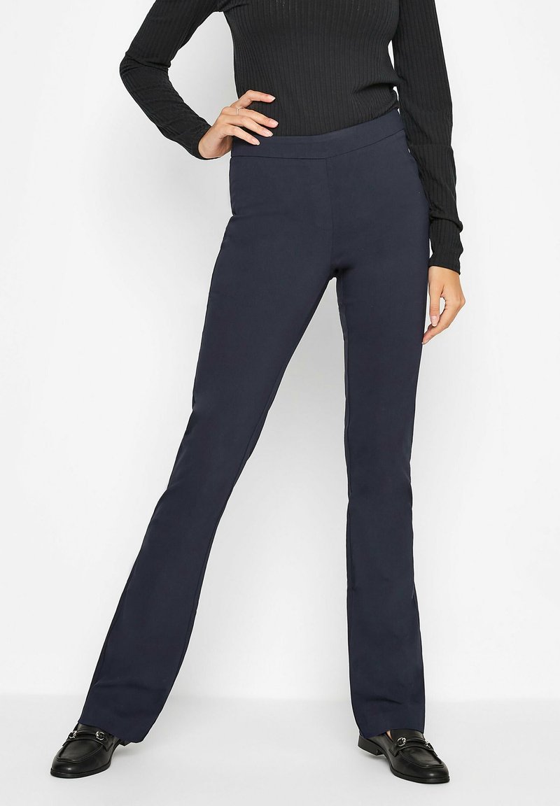 Femme portant un pantalon bleu marine droit, un haut noir côtelé à manches longues et des mocassins noirs, debout avec une main sur la hanche sur fond blanc.