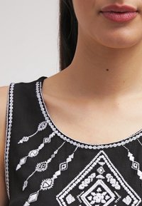 Haut en tissu noir avec une broderie blanche complexe aux motifs géométriques. Encolure dégagée, design sans manches et détails texturés.