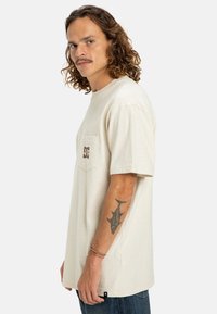 Béžové bavlněné tričko s krátkým rukávem, se zapínací kapsičkou na levém prsu a hnědým logem. Volný střih s kulatým výstřihem.