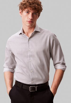 Młody mężczyzna o kręconych włosach, ubrany w dopasowaną, białą koszulę w pionowe paski z podwiniętymi rękawami oraz czarne spodnie z paskiem.