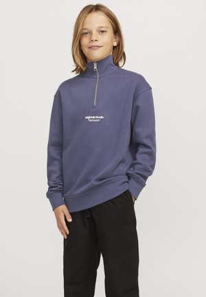 Sudadera de color púrpura oscuro, con un cuello alto y cremallera de un cuarto. En el frente tiene un logo de texto blanco. Se lleva con pantalones negros.